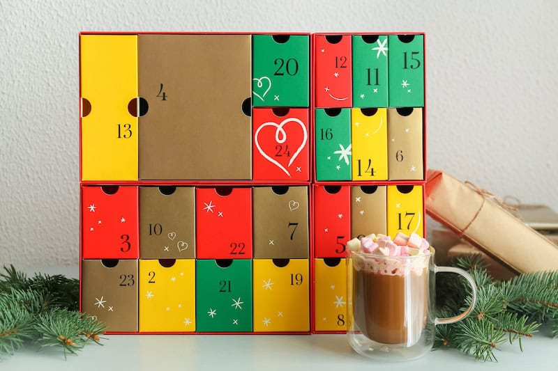 Advent calendar with cup of cocoa and fir branches on shelf near white wall