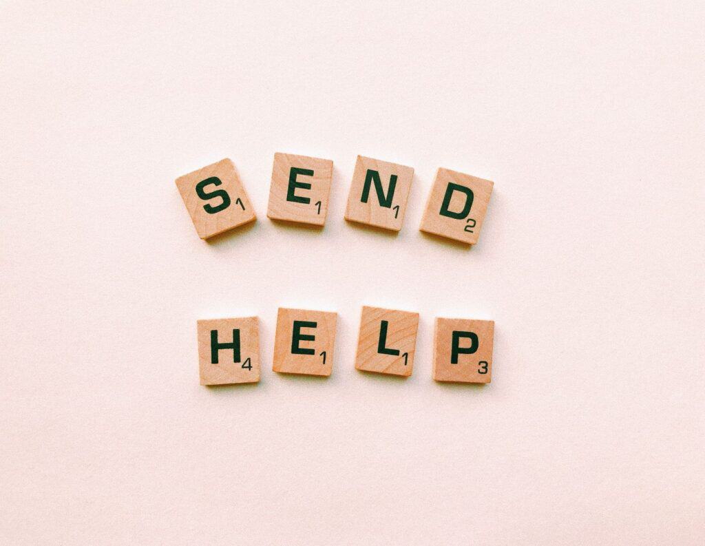 Wooden Scrabble tiles arranged to form the phrase 'send help' on a light pink background.