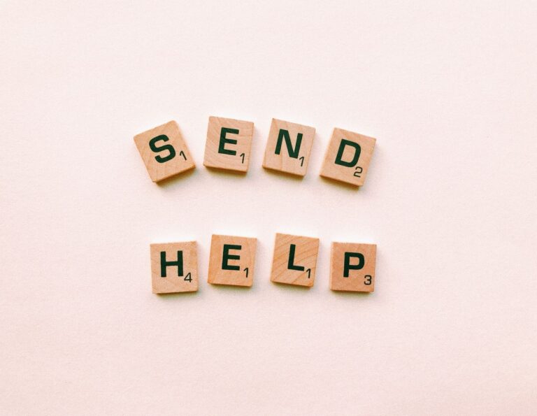 Wooden Scrabble tiles arranged to form the phrase 'send help' on a light pink background.