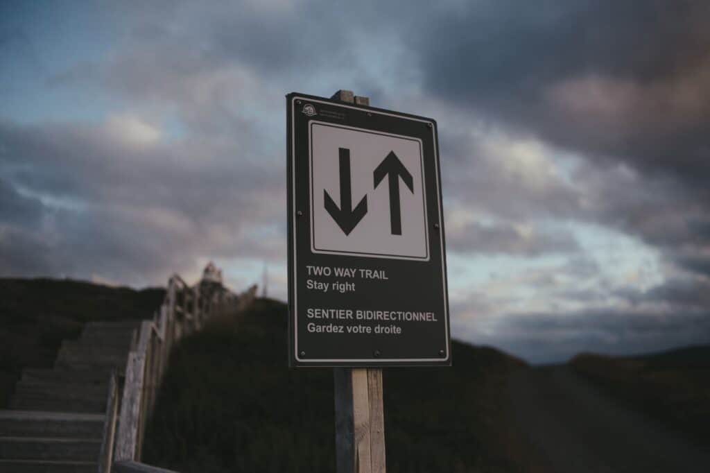 Close-up of a bilingual two-way trail sign against a cloudy sky, outdoors setting.