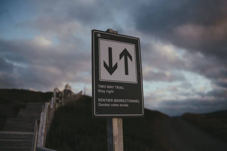 Close-up of a bilingual two-way trail sign against a cloudy sky, outdoors setting.
